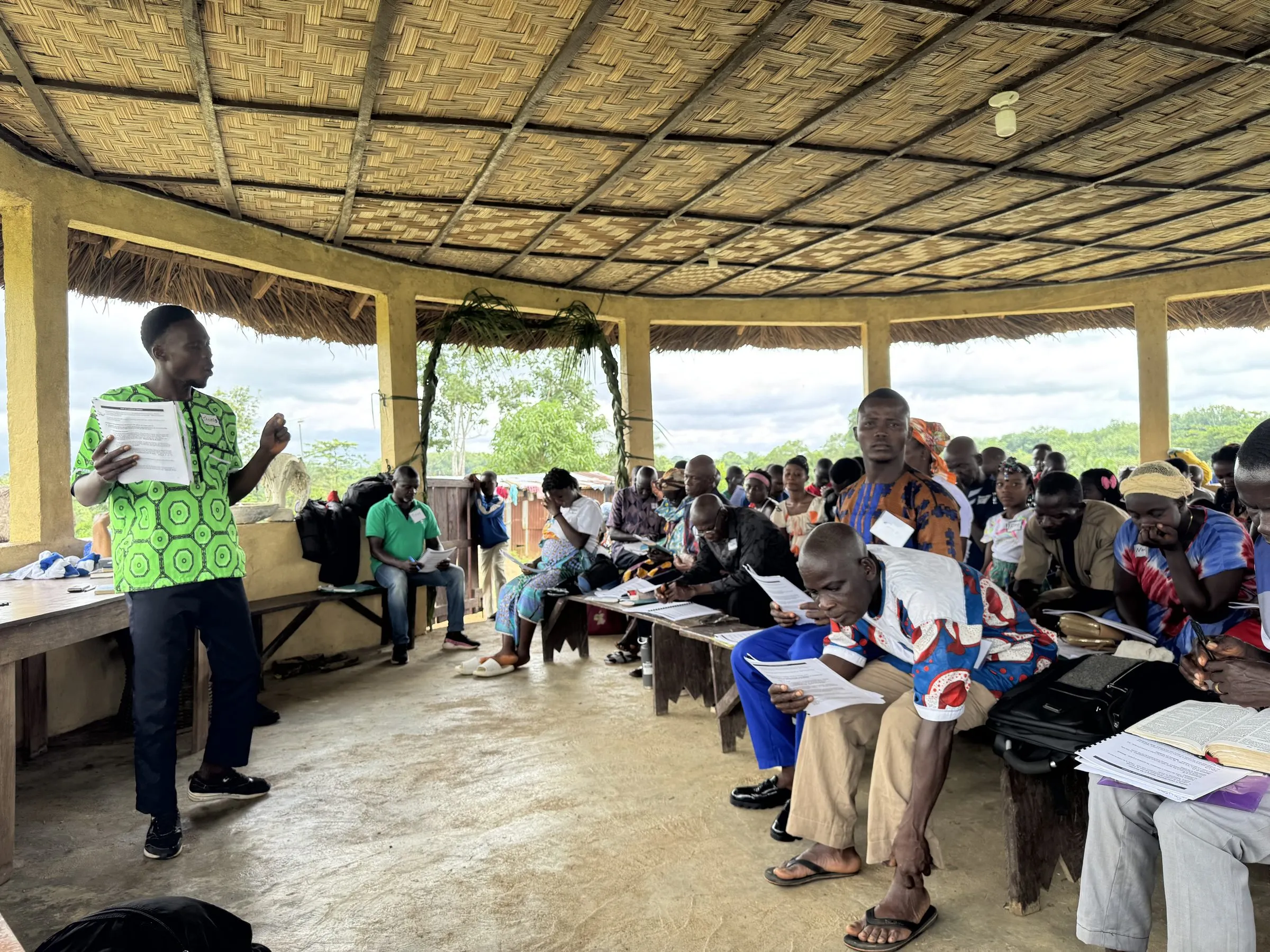 A man teaching from a printed handout to about thirty people seated on benches under a thatched open-air roof, several following along in their own copies.