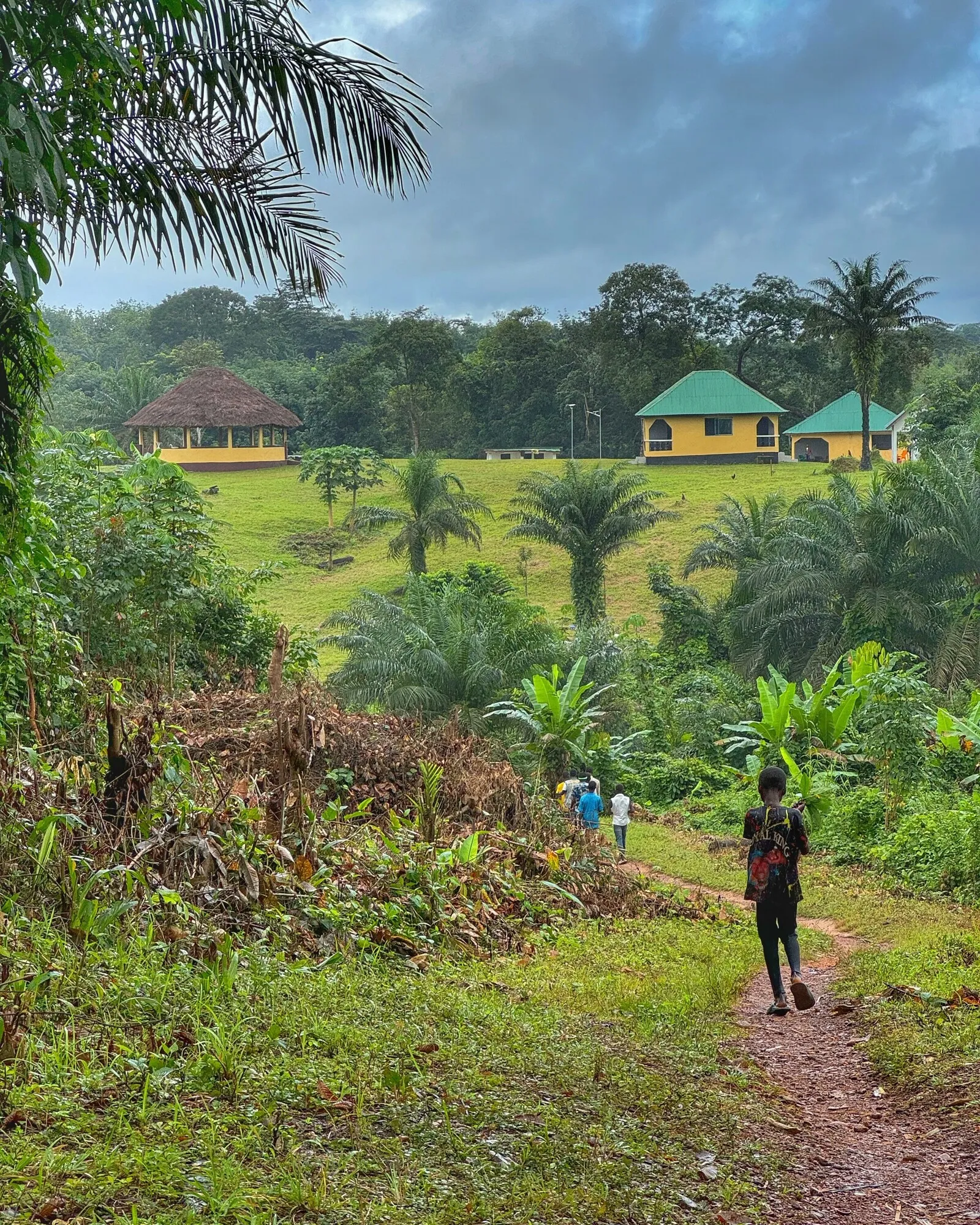 A green hillside in Liberia seen from a red-earth footpath: a yellow building with a green metal roof and a round thatched pavilion stand on the ridge above mown grass, with forest and oil palms all around and people walking up the path.